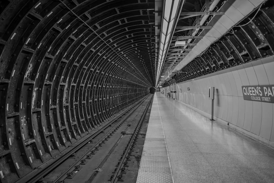 A black and white photograph of Queens Park Station on College Street, showcasing the underground metro tunnel. The left side features a curved, metallic tunnel wall with a repeating pattern of reinfo